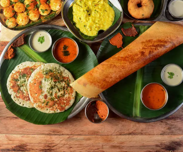 Photo of Assorted South indian breakfast foods on wooden background. Ghee dosa, uttappam,medhu vada,pongal,podi idly and chutney.. Dishes and appetizers of indian cuisine