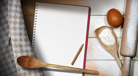 Top view of an empty cookery book on a wooden table with flour, wooden rolling pin, spoons, chicken egg, and brown and white checkered tablecloth. Template for recipes or ingredients list.