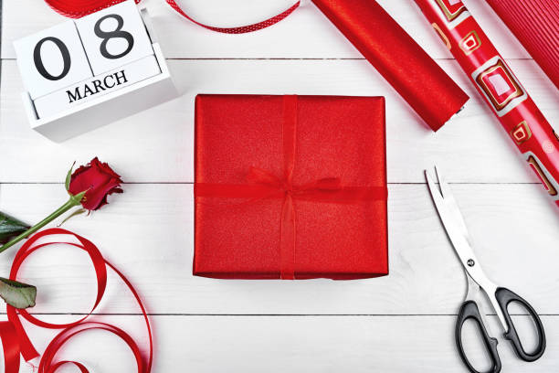istock Gift wrapping background, copy space. Rolls of red paper, ribbon, gift box, rose, scissors and wooden calendar March 8 on wooden background, free space. Flatlay, top view. Greeting card for Womens Day 1292568264