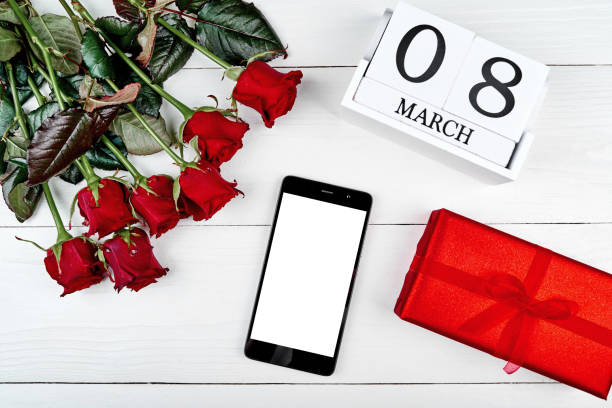 Red gift boxes, roses, cellphone with blank screen and cube calendar on white wooden background, copy space. Save the date, Womens Day, March 8. Greeting card. Top view, flat lay