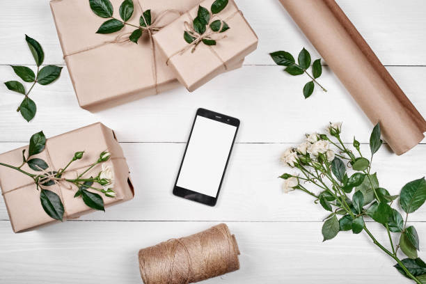 istock Gift boxes wrapped in kraft with fresh roses and mobile smartphone with white screen on wooden background, copy space. Greeting card mockup. Top view, flat lay. Spring, Mothers day, Womans Day 1292568354