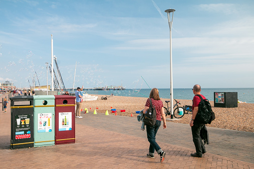 People on Brighton beach, strolling and enjoying the sun. In the background a father holds his child and bubbles float through the air and in the foreground are dustbins with images depicting what should be thrown where.