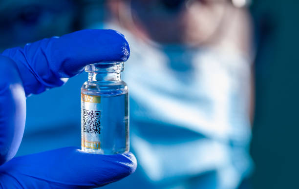 Vial with vaccine injection held by healthcare worker's hand in blue surgical protective glove, with face of the assistant covered by protective blue surgical mask in the background stock photo