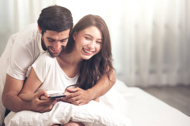 casal usando telefone celular na cama branca no tempo da manhã - boyfriend heterosexual couple married activity - fotografias e filmes do acervo
