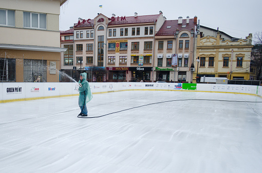 Ivano-Frankivsk, Ukraine - November, 2019: Man in raincoat filling ice rink with water from hose. City public ice rink near.