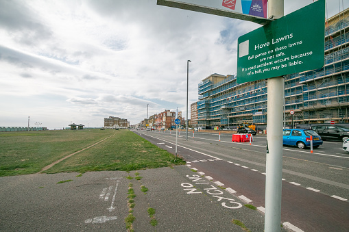 A no cycling road marking on Hove Lawns in Brighton & Hove, England, with cars visible on the road and people in the far distance. A sign warns people that ball games are dangerous as they can cause road accidents, leaving the person who caused the ball to go into the traffic liable to prosecution.