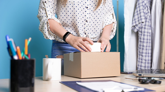 Small business, young female entrepreneur at the table packing a small box of goods, parcel for online store customers. Workplace, portrait on a blue background