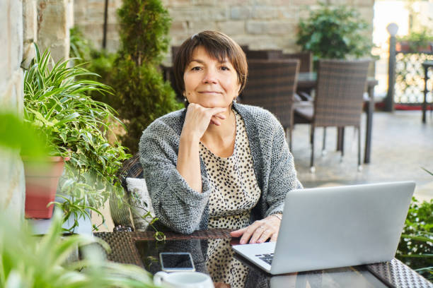 verticale de femme d’affaires adulte avec l’ordinateur portatif à la table dans le café d’été, travaillant dehors et souriant à l’appareil-photo. concept indépendant de femme âgée de pigiste - business slave photos et images de collection