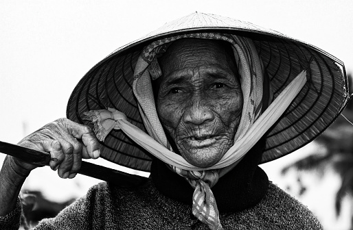 Hoi-An, Vietnam - December 9. 2001: Portrait of old poor farmer woman with traditional conical rice hat carrying heavy loads with stick