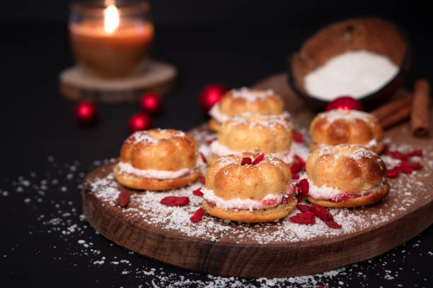 Goji berries and red shiny Christmas balls. Coconut shell full of flour, sprinkled over five small home baked cakes, filled with strawberry cream. Wooden board. Cinnamon sticks decoration. Burning candle. Black background bayas de goji y bolas de navidad rojas brillantes. concha de coco llena de harina, espolvoreada sobre cinco pequeños pasteles caseros horneados, rellenos de crema de fresa. tablero de madera. decoración de palitos de canela. vela encendida. fondo negro