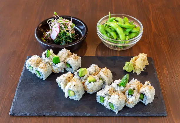 Photo of Japanese sushi food with avocado, rice, sea delicacies on a black stone, on a brown background. Plates with salad and green beans.