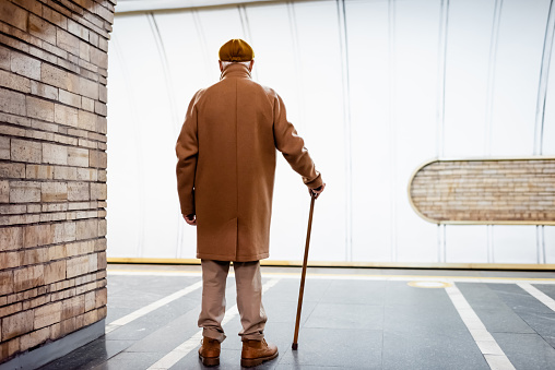back view of aged man in autumn outfit standing with walking stick on underground platform