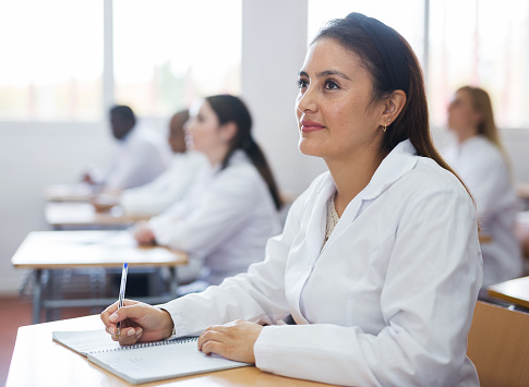 Positive latin american girl in white coat listening to lecture and taking notes in classroom during professional training