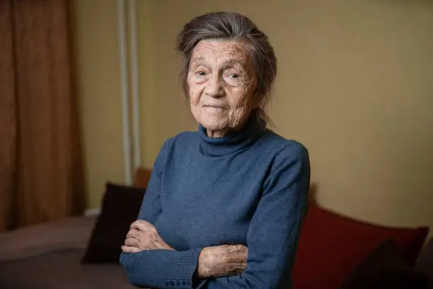 Elderly Caucasian senior grandmother ninety years old looks attentively and smiles, feels happy, large portrait, face with deep wrinkles, gray hair. Theme pensioners, old people and long-lived woman.