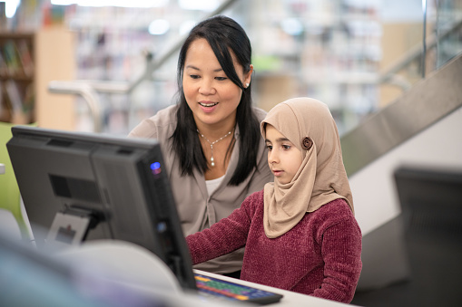 A Muslim girl is getting some help with her computer from her Asian teacher. They are at a public library.