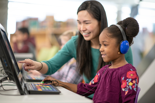 An African American girl is getting some help from her teacher with her computer. They both appear to be at their school library.