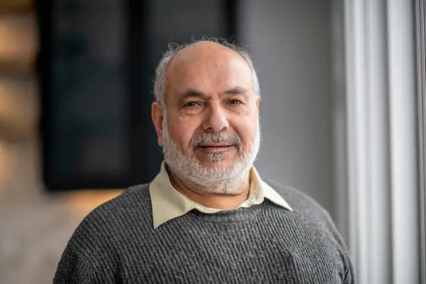 An elderly man of Middle Eastern ethnicity is looking at the camera while wearing a grey sweater. He is standing next to big bright windows at his home.