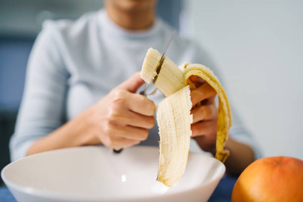 Caucasian woman sitting by the table at home cutting banana - Adult girl female preparing fruit salad at home - healthy eating concept copy space close up stock photo