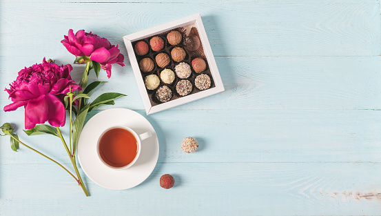 Cup of tea box of chocolates burgundy beautiful peonies on blue gentle background. Selective focus copy space. Top view