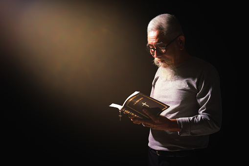 christianity background of asian senior man standing in dark background holding and reading holy bible with light ray from above