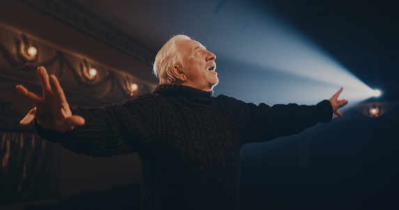 Tracking shot of aged man in sweater speaking and gesticulating while turning around in spotlight on stage during performance in dark theater
