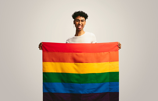 Man sticking out tongue and winking holding pride flag. Gay man with lgbt flag making funny face on white background.