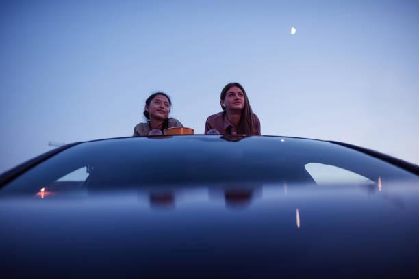 é hora do filme. duas garotas deitadas no telhado de um carro com sacos de pipoca e bebidas enquanto assistiam a um filme em um cinema ao ar livre à noite - outdoors drinking women friendship - fotografias e filmes do acervo