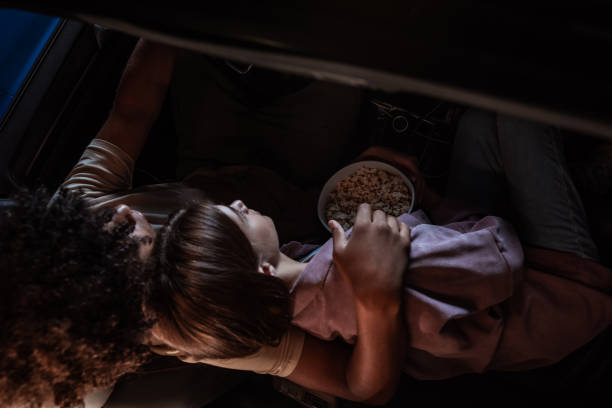 top view of a young couple watching a movie, having popcorn while sitting together in the car parked in front of a big screen in an open air cinema - drive in movie car inside of billboard imagens e fotografias de stock