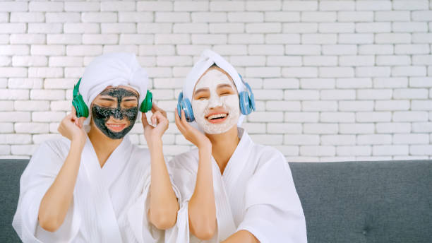 dwie szczęśliwe azjatyckie dziewczyny w białych szlafrokach z maską na twarz, które cieszą się muzyką ze słuchawkami w salonie. - moisturizing cream audio zdjęcia i obrazy z banku zdjęć
