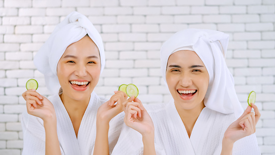 Two happy Asian girls in white bathrobes with towels on heads holding cucumber slices in living room.