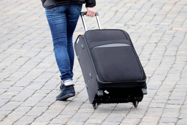 femme dans des jeans marchant avec une valise sur des roues - valise à roulettes photos et images de collection