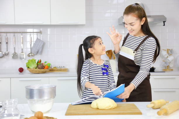 feliz madre asiática enseñando a su hija pequeña en la pan de hornear de la masa dentro de la cocina moderna blanca mientras mira el libro de cocina - bun bread 7 grain bread dough fotografías e imágenes de stock