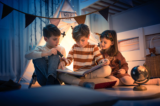 Two boys and a girl reading a fairy tale book with flashlight at night camping play in kids room