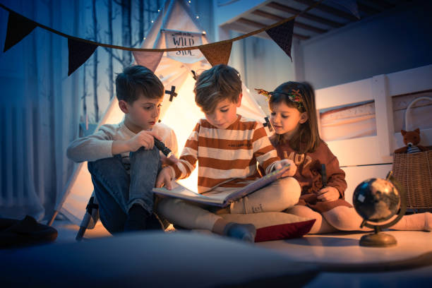 Cute kids play camping in kids room Two boys and a girl reading a fairy tale book with flashlight at night camping play in kids room bedtime photos stock pictures, royalty-free photos & images