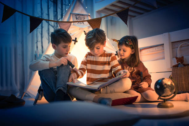 Cute kids play camping in kids room Two boys and a girl reading a fairy tale book with flashlight at night camping play in kids room boy and girl reading stock pictures, royalty-free photos & images