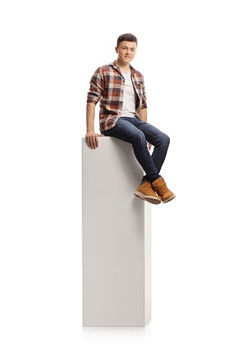 Teenag guy sitting on a white column and smiling at camera isolated on white background