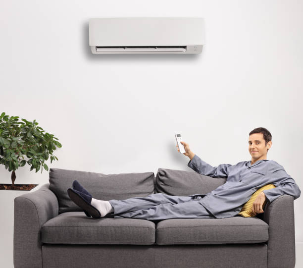 istock Man in pajamas resting on a gray sofa and holding a remote control from an air conditioning unit 1292732897