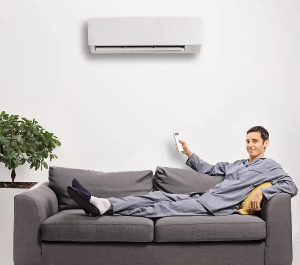 Photo of Man in pajamas resting on a gray sofa and holding a remote control from an air conditioning unit