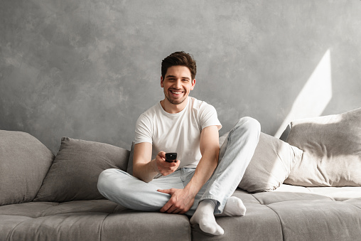 Photo of joyful man 30s in casual clothing sitting on sofa in living room and looking on camera with remote control in hand