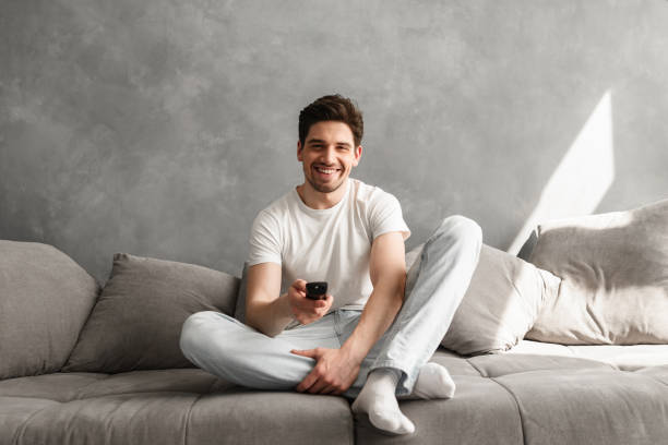 Photo of joyful man 30s in casual clothing sitting on sofa in living room, and looking on camera with remote control in hand stock photo