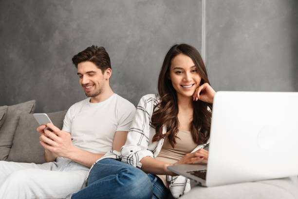 istock Joyful man and woman sitting together on couch in gray interior, while using laptop and smartphone 1292734703