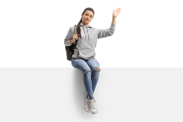 istock Cheerful female student sitting on a blank board and waving at camera 1292735225