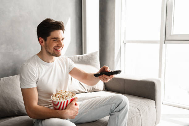 istock Handsome man sitting on sofa at home and changing TV channels, while holding pop corn 1292736471