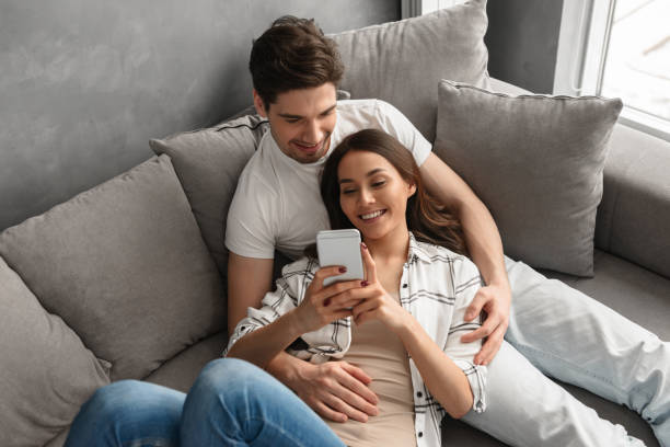 Homme et femme caucasiens de couples dans l’habillement occasionnel se reposant dans le salon à la maison, et regardant le smartphone - Photo