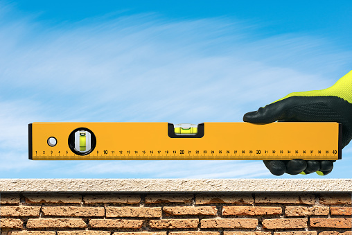 Closeup of a hand with work glove, holding an orange construction spirit level, over a brick and marble wall with blue sky on background.