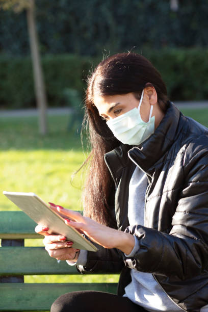 istock Young woman with face mask sitting on a bench in a public park using digital tablet 1292736908
