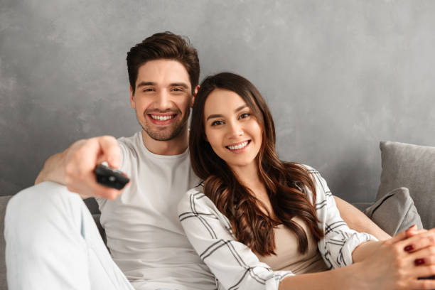 image closeup of young family sitting on sofa at home and looking at you, showing remote control at camera - apartment television family couple imagens e fotografias de stock