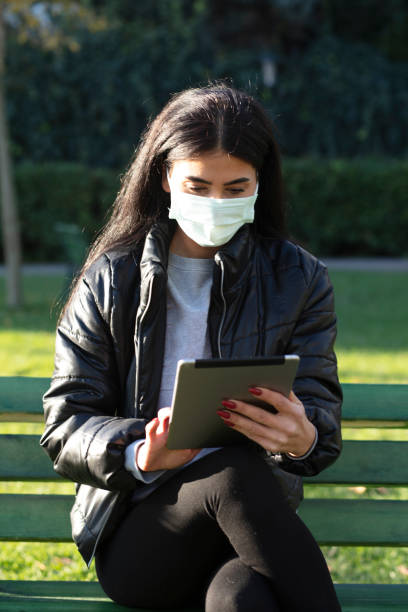istock Young woman with face mask sitting on a bench in a public park using digital tablet 1292737929