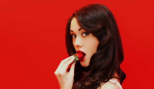 Portrait close up of attractive brunette sexy woman enjoying a taste of fresh strawberry on a colorful red background stock photo