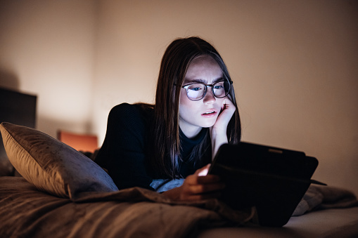 Angry, worried and disappointed looking teenage woman lying on her bed in bedroom at night relaxing. Reading E-Mails, Chat Messages and Social Media Postings on her Digital Tablet Computer Ambient Bedroom Night Light. Millenial Generation Modern Technology Lifestyle.